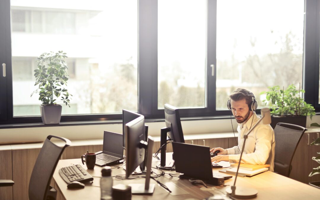 A businessman sits at a desk using multiple computers and a headset in a well-lit modern office.